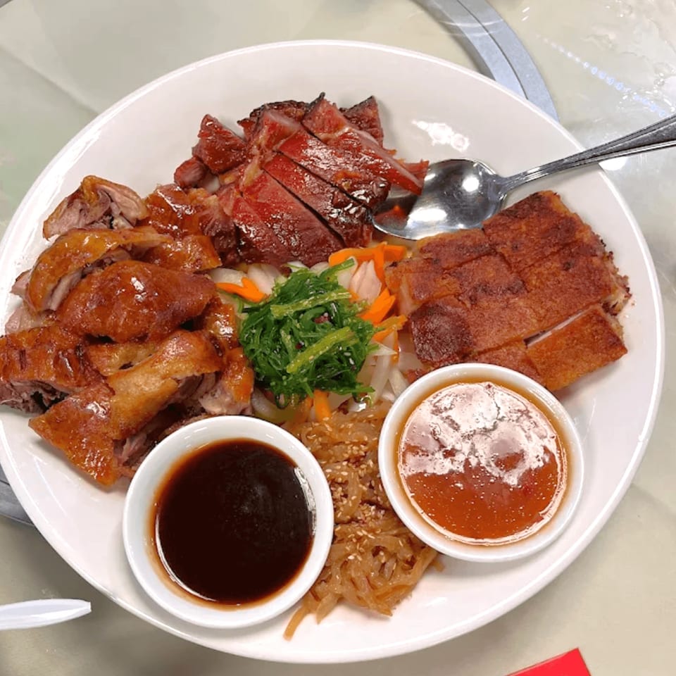 A white plate with sliced roasted duck, char siu, crispy pork belly, seaweed salad, and two dipping sauces.